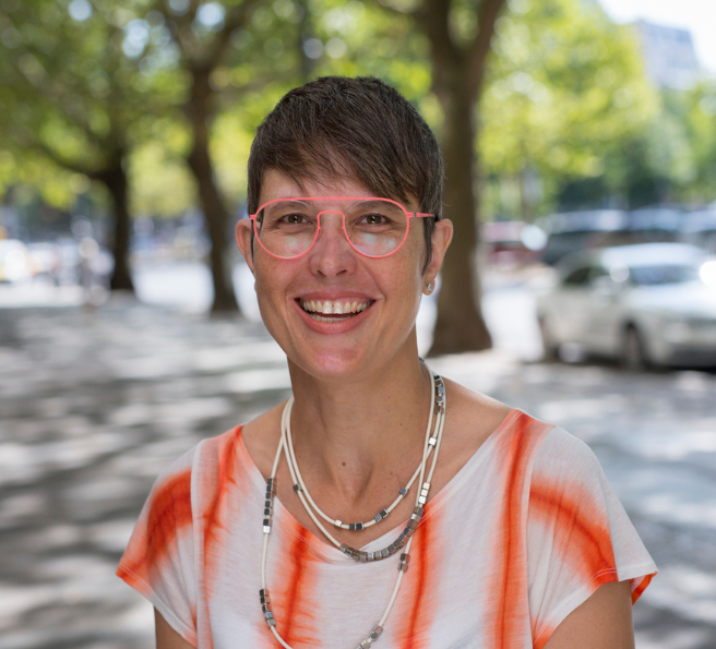Coach Fabienne Cuisinier wearing an orange-white shirt, orange glasses and necklaces, smiling into the camera. In the summerly background are a street and trees.