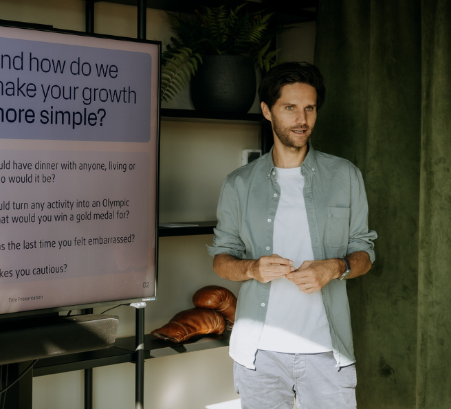 A man in a business casual shirt is presenting something in front of a green curtain and a big screen