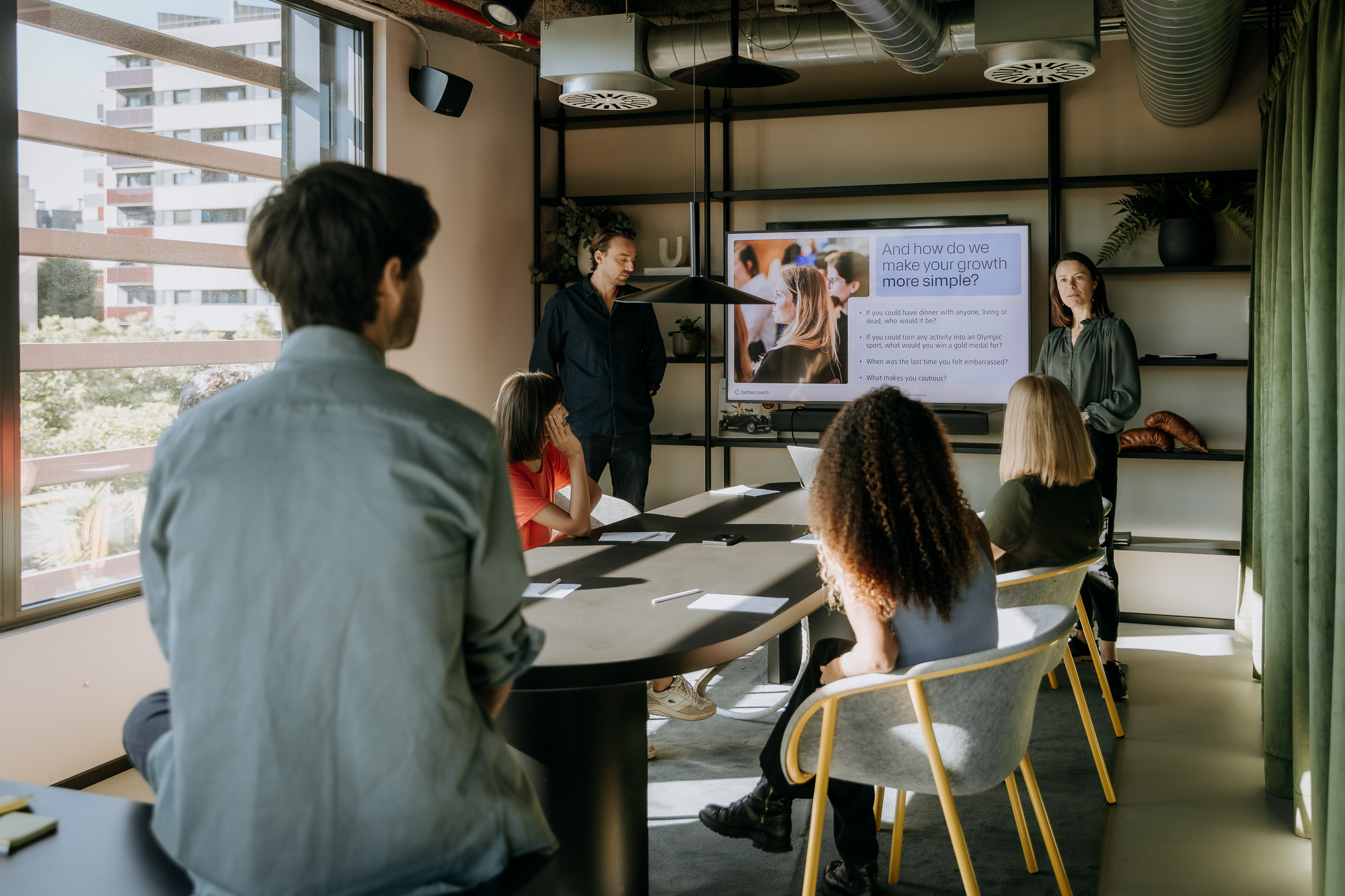 6 people are in a room, two are presenting something besides a screen, 3 people are sitting around a table and one person is with their back to the camera sitting on a shelf in the front, listening to the presentation